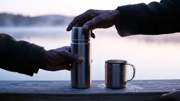 A weathered hand pouring black coffee from a steel thermos into a cup on a dock rail at dawn