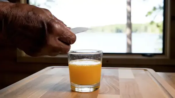 A hand tearing the corner of a small white gel pouch and squeezing the gel into a glass of orange juice