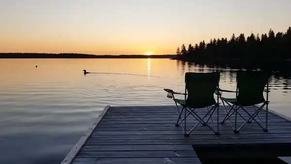 An empty dock with two folding chairs on a still lake at sunset, the light shimmering on the water